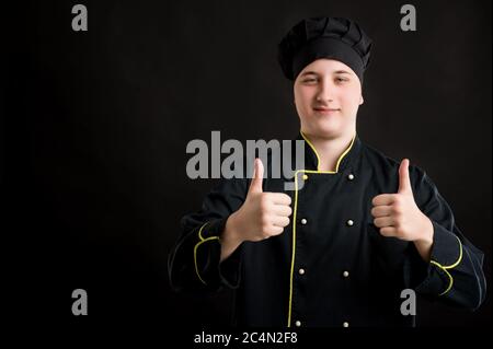 Portrait eines jungen Mannes in einem schwarzen Koch Anzug gekleidet zeigt Daumen nach oben mit beiden Händen posiert auf einem schwarzen isolierten Hintergrund mit Copy Space advertisin Stockfoto