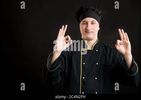 Portrait von jungen Mann in einem schwarzen Koch Anzug gekleidet zeigt Doppel OK-Zeichen posiert auf einem schwarzen isolierten Hintergrund mit Kopie Raum Werbefläche Stockfoto