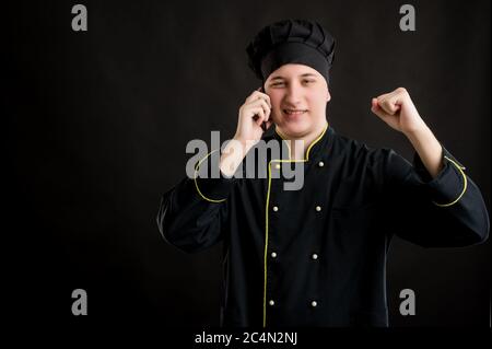 Porträt eines jungen Mannes in einem schwarzen Koch Anzug mit gekleidet, glückliches Gesicht und Gesten und sprechen auf Handy, posiert auf einem schwarzen isolierten Hintergrund. Stockfoto