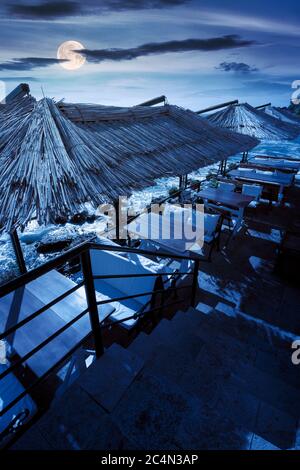 Terrasse eines Restaurants am Meer in der Nacht. Schöne Aussicht in die Bucht im Vollmondlicht. Dekorativer Schirm über Holztischen und Sitzplätzen Stockfoto