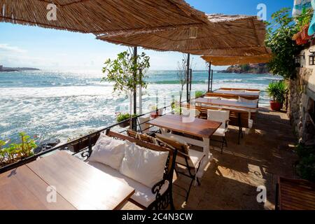 Leere Terrasse eines Restaurants am Meer. Schöner Blick in die Bucht im Morgenlicht. Dekorativer Schirm über Holztischen und Sitzplätzen. Sonniges Wetter Stockfoto