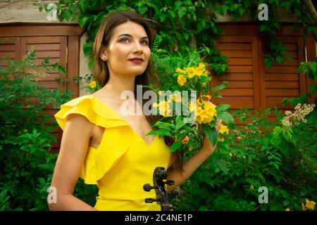 Ein Porträt eines schönen jungen Musikermädchen in gelbem Kleid mit einer Geige in der Hand. Grüne Pflanzen, gelbe Blumen und hölzerne Fensterläden auf der Rückseite Stockfoto