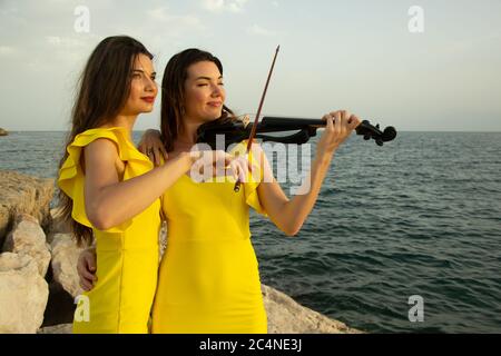 Zwei schöne Mädchen Geiger in gelben Konzertkleider spielen elektrische Geigen mit Sonnenuntergang, Bergen und Mittelmeer im Hintergrund. Stockfoto