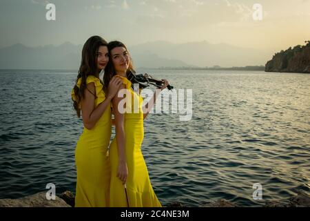 Zwei schöne Mädchen Geiger in gelben Konzertkleider spielen elektrische Geigen mit Sonnenuntergang, Bergen und Mittelmeer im Hintergrund. Stockfoto