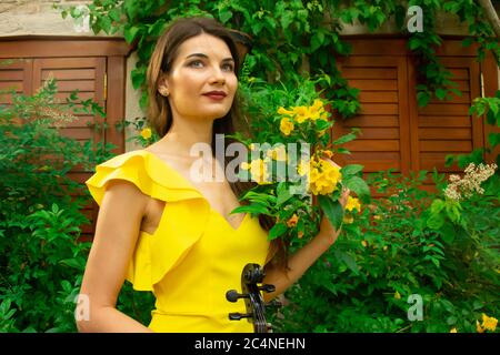 Ein Porträt eines schönen jungen Musikermädchen in gelbem Kleid mit einer Geige in der Hand. Grüne Pflanzen, gelbe Blumen und hölzerne Fensterläden auf der Rückseite Stockfoto