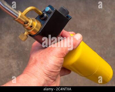 Nahaufnahme POV Aufnahme einer Hand eines Mannes mit einer traditionellen Lötlampe / Taschenlampe, mit einem leuchtend gelben Gasflasche / Kanister. Stockfoto