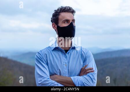 Junger Mann trägt schwarze Maske zuversichtlich in die Zukunft schauen blau Button up Shirt blaue Augen dunkelbraune Haare gute Knochenstruktur gekreuzte Arme reich Stockfoto