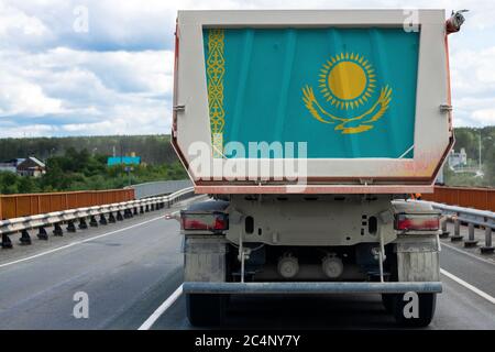 Großer schmutziger LKW mit der Nationalflagge von Kasachstan bewegt sich auf der Autobahn, vor dem Hintergrund des Dorfes und Waldlandschaft. Konzept der Exp Stockfoto