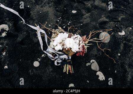 brautstrauß aus weißen Rosen, Lilie, Burgunder Nelken, protea, getrockneten Zweigen, Brunia und Leukadendron mit grauen und schwarzen Bändern auf dem schwarzen Sand in Stockfoto