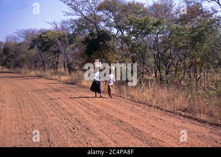 Zwei junge Frauen, die auf einer unbefestigten Straße mit Lasten auf dem Kopf spazieren, Hwange National Park, Simbabwe Stockfoto