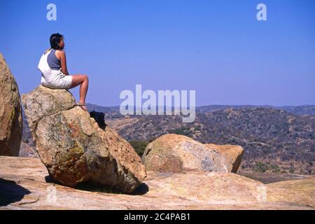 Junge Frau, die auf einem großen Felsblock sitzt und den Blick auf World's View, Matobo National Park, Simbabwe, in der Nähe des Grabes von Cecil Rhodes bewundert Stockfoto