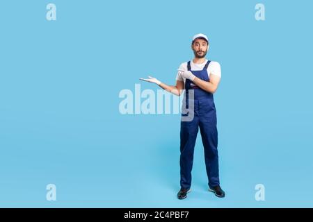 In voller Länge überrascht Arbeiter in blauen Overalls, Kappe und Schutzhandschuhe stehen, zeigen leeren Raum auf seinem Arm und Blick mit schockiert erstaunt ex Stockfoto