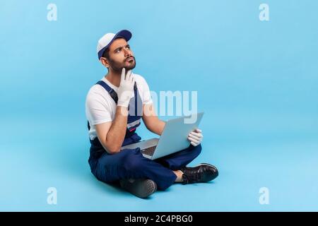 Reparaturservice, Online-Hilfe. Portrait des pensive Handwerker in Arbeitskleidung sitzen mit gekreuzten Beinen mit Laptop, über die Entscheidung denken, nicht sicher, Zweifel haben Stockfoto