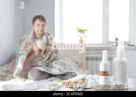 Ein kranker Kerl sitzt zu Hause auf der Couch mit einem Taschentuch und nimmt Medizin. Es gibt ein Thermometer, Nasentropfen und Pillen auf dem Nachttisch Stockfoto