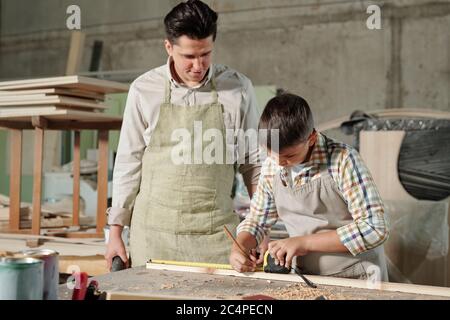 Zimmermann mittleren Alters in Schürze am Schreibtisch stehend und beobachten, wie sein Teenager-Sohn auf Plank in der Werkstatt Zeichen machen Stockfoto