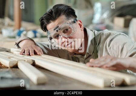 Konzentrierter männlicher Zimmermann mittleren Alters in Schutzbrille, der die Oberfläche der Holzbohlen in der Werkstatt untersucht Stockfoto