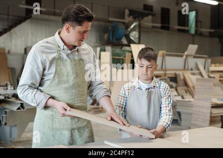 Geschickter Zimmermann in Schürze berühren Holz, während der Sohn in der Möbelwerkstatt bearbeitet Plank Stockfoto