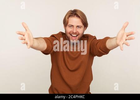Lasst mich Euch umarmen! Portrait von glücklich fröhlich gastfreundlich bärtigen Mann in Sweatshirt stehend mit ausgestreckten Händen, lächelnd und einladend mit freien hu Stockfoto