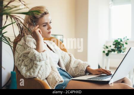 Nachdenkliche kaukasische Frau mit blonden Haaren mit einem Laptop zu Hause trägt einen warmen Strickpullover Stockfoto