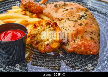 Nahaufnahme von gebackenen Hühnerschenkeln mit pommes Frites, gegrilltem Mais und Ketchup auf dunklem Holztisch Stockfoto