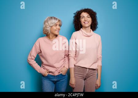 Charmante Frau mit lockigem Haar und ihre Freundin mit brünettem Lächeln in der gleichen Kleidung auf einer blauen Studiowand Stockfoto