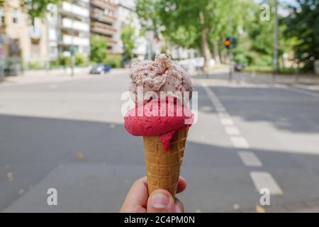 Ausgewählte Fokusansicht auf der Hand halten 2 Kugeln Schokolade Chip und Himbeere auf Waffel-Kegel mit verschwommenem Hintergrund der sonnigen Straße in der Sommersaison. Stockfoto
