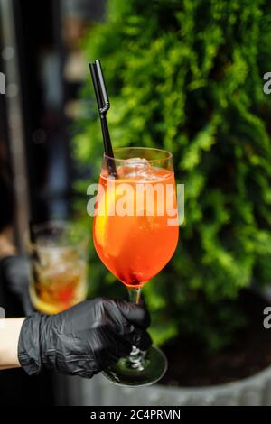 Hell und bunt Cocktail aperol Spritz in einer Kellnerhand in einem Handschuh vor einem Hintergrund von Grün Stockfoto