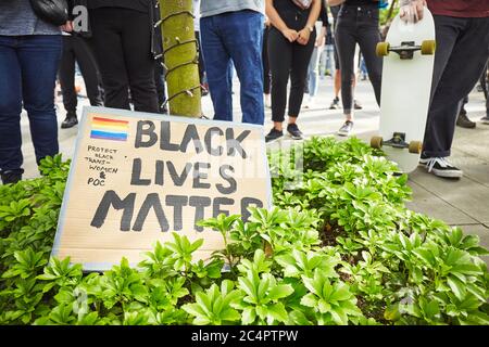 Ein Pappschild, das auf einem Baum mit einem kleinen Stolz Regenbogen lehnt und geschrieben "schwarze Leben Materie, Menschen stehen herum Stockfoto