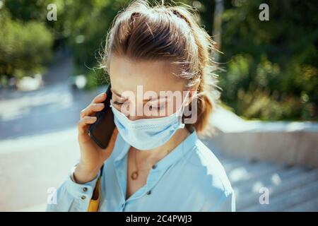 Leben während covid-19 Pandemie. Junge Frau in blauer Bluse mit medizinischer Maske sprechen auf einem Smartphone im Freien in der Stadt. Stockfoto