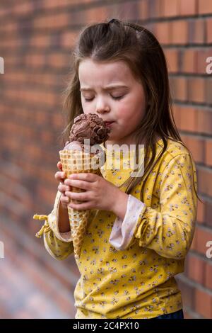 Nahaufnahme eines entzückenden kleinen Mädchen essen eine große Schokolade Eis Kegel mit einer Ziegelwand im Hintergrund Stockfoto