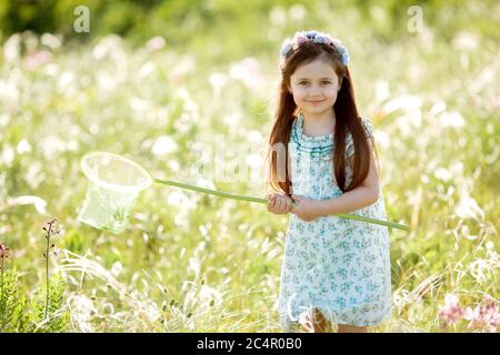 Nettes kleines Mädchen mit einem Kranz auf dem Kopf geht in einem Feld und fängt Schmetterlinge Netz Stockfoto