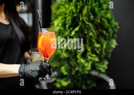 Hell und bunt Cocktail aperol Spritz in einer Kellnerhand in einem Handschuh vor einem Hintergrund von Grün Stockfoto