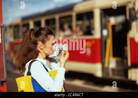 Leben während der Coronavirus-Pandemie. Elegante Frau in blauer Bluse mit Handtasche in medizinischer Maske an der Straßenbahnhaltestelle im Freien auf der Straße der Stadt. Stockfoto