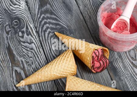 Erdbeereis in einem Waffelkegel. Garniert mit Schokolade. In der Nähe gibt es leere Waffelkegel ohne Eis. Eisbehälter und Löffel. Auf pi Stockfoto