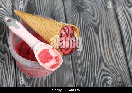 Erdbeereis in einem Waffelkegel. Garniert mit Schokolade. In der Nähe des Behälters mit Eis und einem Löffel dafür. Auf Pinien-Brettern in schwarz lackiert Stockfoto