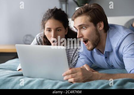 Schockiert junges Paar auf Laptop-Bildschirm, auf dem Bett liegend Stockfoto