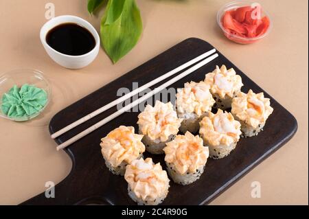 Brötchen mit Sahne-Sauce und gebratenen Garnelen gekrönt. Sushi-Brötchen auf dunklem Holzbrett mit Wasabi, Ingwer und Essstäbchen. Beigefarbener Hintergrund mit Stockfoto