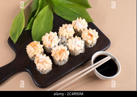 Brötchen mit gelber Sahne-Sauce und gebratenen Garnelen gekrönt. Sushi-Rollen auf dunklem Holzbrett. Beige Hintergrund mit Blättern Stockfoto