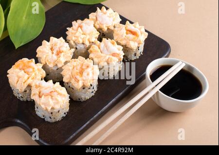 Brötchen mit Sahne-Sauce und gebratenen Garnelen gekrönt. Sushi-Rollen auf dunklem Holzbrett. Beigefarbener Hintergrund Stockfoto