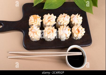 Brötchen mit Sahne-Sauce und gebratenen Garnelen gekrönt. Sushi-Rollen auf dunklem Holzbrett. Beigefarbener Hintergrund Stockfoto