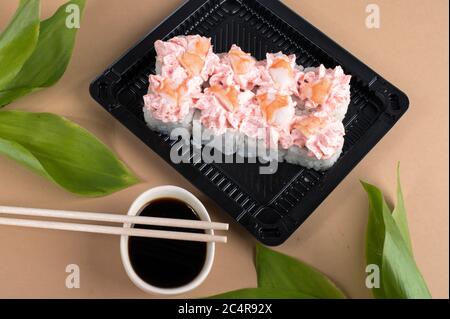Sushi-Brötchen mit rosa Frischkäse und Garnelen gekrönt. Sushi-Rollen in schwarzen Kunststoffbehältern mit Sojasauce und Essstäbchen gesetzt Stockfoto