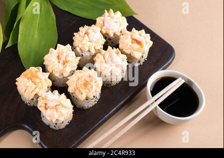 Brötchen mit gelber Sahne-Sauce und gebratenen Garnelen gekrönt. Sushi-Rollen auf dunklem Holzbrett. Beige Hintergrund mit Blättern Stockfoto