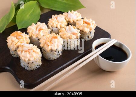 Brötchen mit gelber Sahne-Sauce und gebratenen Garnelen gekrönt. Sushi-Rollen auf dunklem Holzbrett. Beige Hintergrund mit Blättern Stockfoto