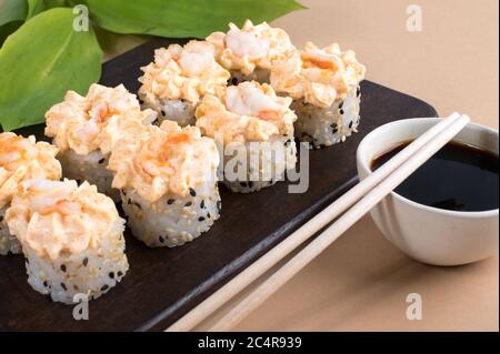 Brötchen mit gelber Sahne-Sauce und gebratenen Garnelen gekrönt. Sushi-Rollen auf dunklem Holzbrett. Beige Hintergrund mit Blättern Stockfoto