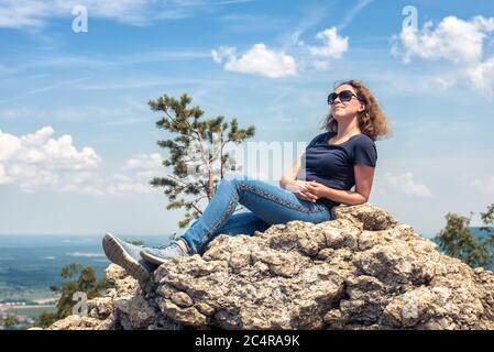Frau mit Sonnenbrille, die im Sommer auf dem Berggipfel entspannt. Landschaft und glücklich schöne Person Sonnenbaden auf Klippe. Erwachsene hübsches Mädchen Ruhe nach Clim Stockfoto