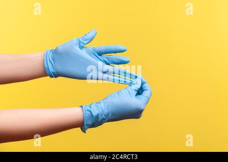 Profil Seitenansicht Nahaufnahme der menschlichen Hand in blauen OP-Handschuhen zeigt, wie man Handschuhe nimmt. Indoor, Studio-Aufnahme, isoliert auf gelbem Hintergrund. Stockfoto