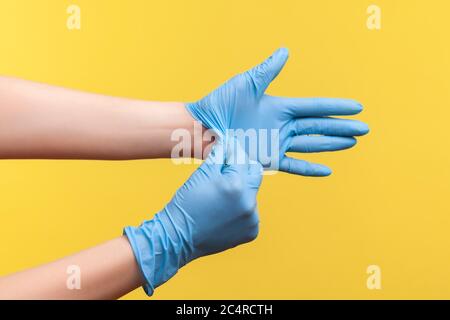 Profil Seitenansicht Nahaufnahme der menschlichen Hand in blauen OP-Handschuhen zeigt, wie man Handschuhe nimmt. Indoor, Studio-Aufnahme, isoliert auf gelbem Hintergrund. Stockfoto