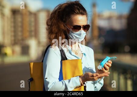 Leben während Coronavirus Pandemie. Junge Frau in blauer Bluse mit medizinischen Maske, Sonnenbrille und Handtasche Desinfektion Hände mit Desinfektionsmittel im Freien in t Stockfoto