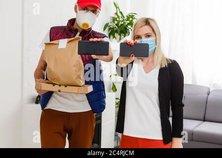 Junge Frau trägt medizinische Maske Empfangen Paket von Lieferung Mann drinnen. Verhinderung der Ausbreitung von Viren Stockfoto