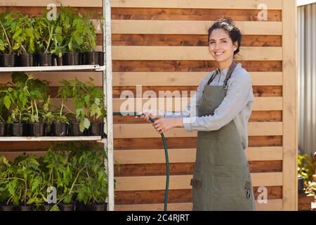 Portrait einer lächelnden Arbeiterin, die Topfpflanzen an einer Holzwand wässern und dabei die Arbeit auf dem Bauernhof genießen, Platz kopieren Stockfoto